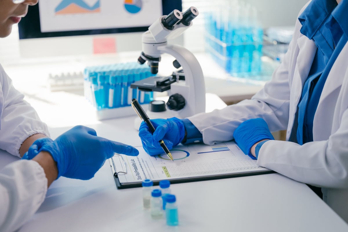 Two scientists wearing lab coats and gloves examining data on a clipboard in a modern laboratory