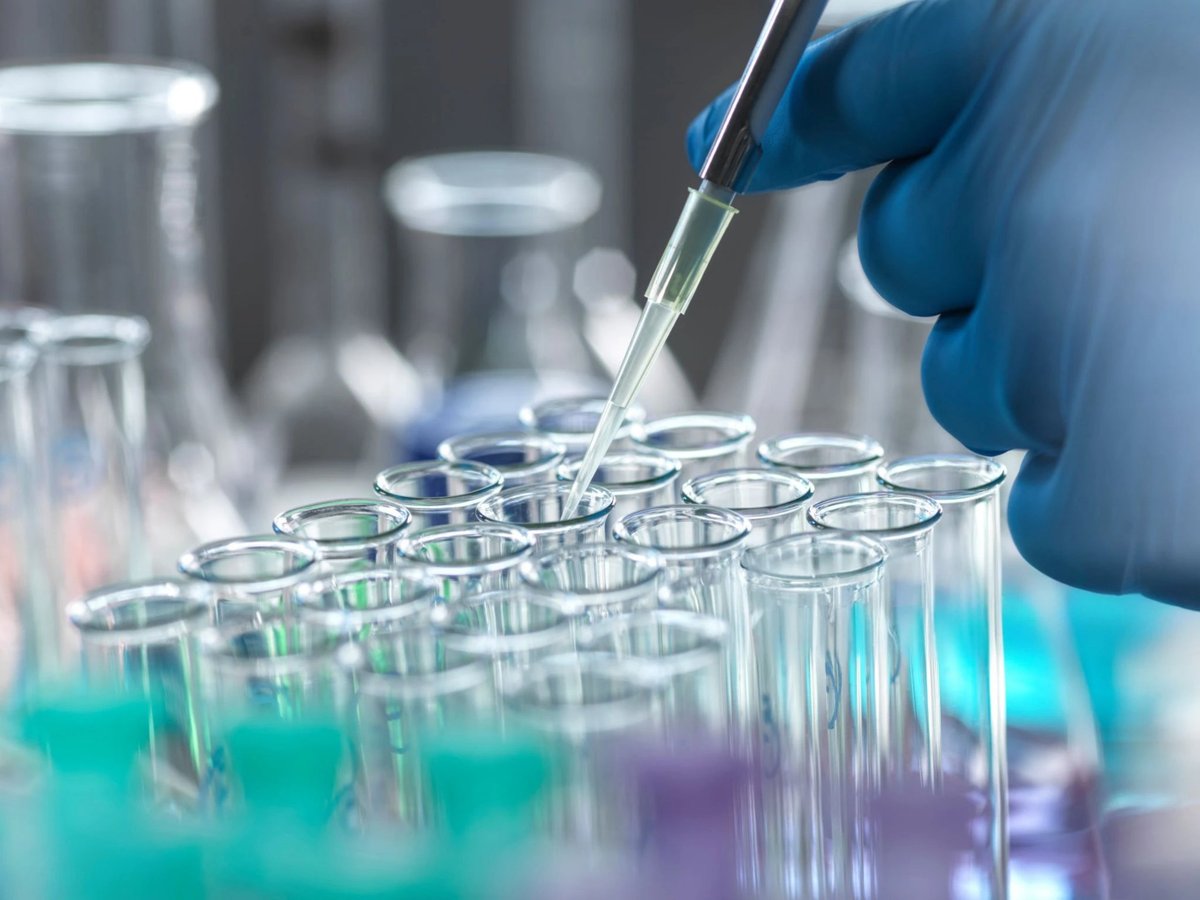 A gloved hand carefully uses a pipette to dispense liquid into a row of laboratory test tubes