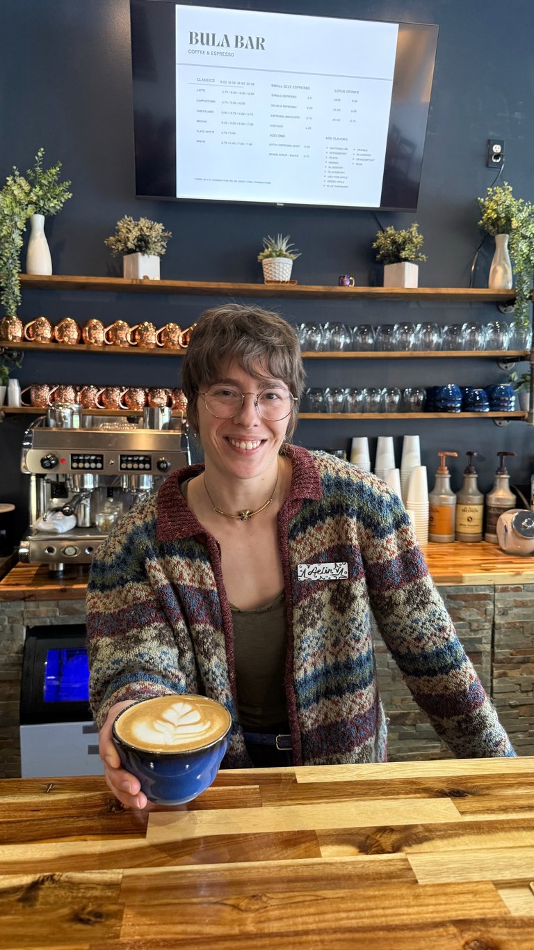 Smiling barista holding a latte with latte art at a coffee bar counter, with menu board and shelves of equipment visible behind