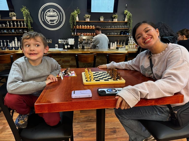 Woman and young boy playing chess at a table in a cafe, both smiling at the camera