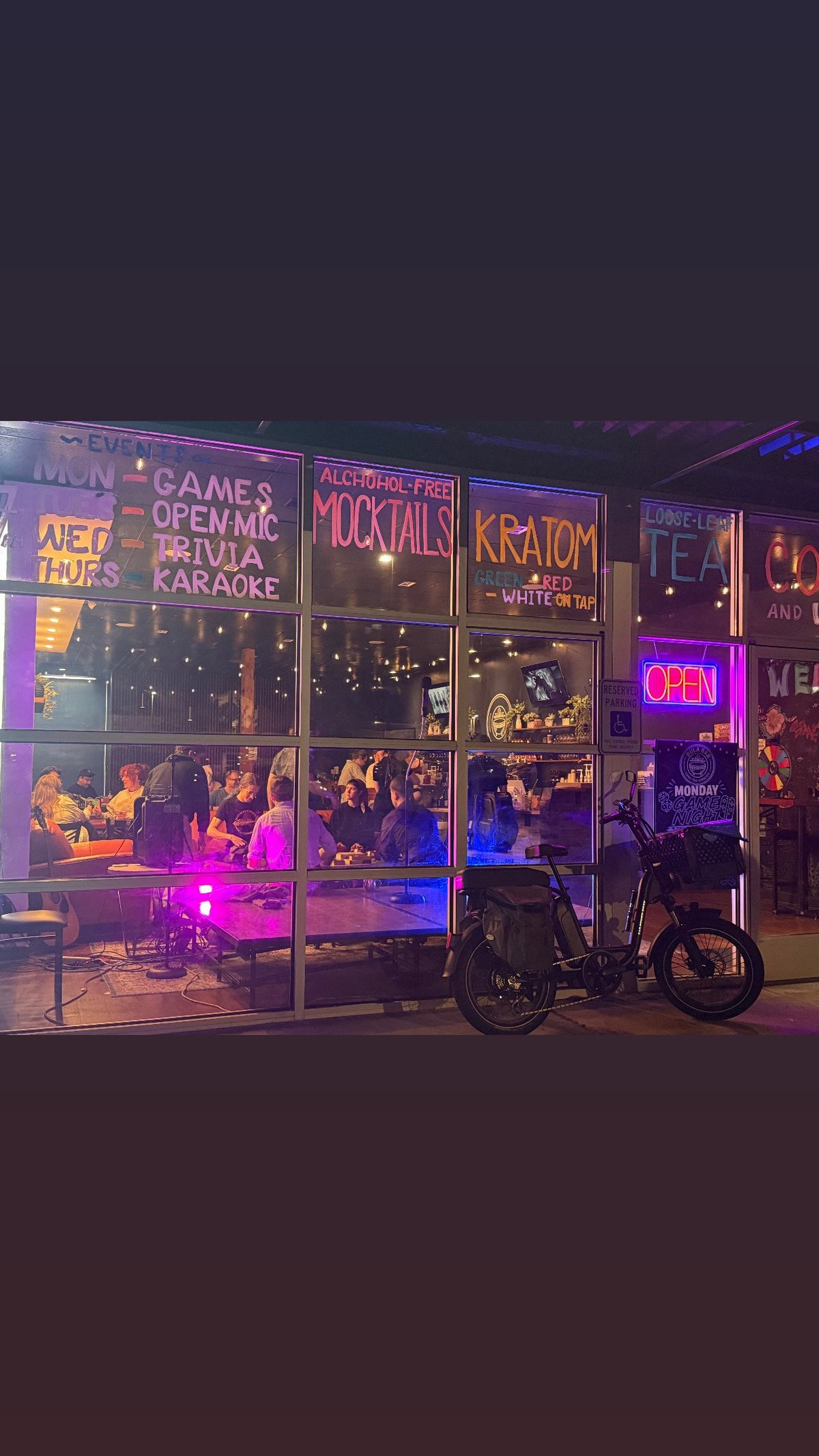 Nighttime storefront with neon signs advertising games, trivia, karaoke and cocktails. A bicycle is parked in front of the illuminated venue entrance.