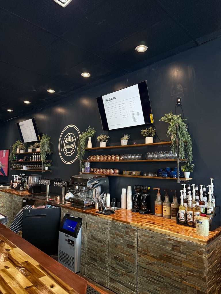 Coffee shop counter with barista, wooden bar, menu display, shelves of beverages, and dark accent wall with plants and lights