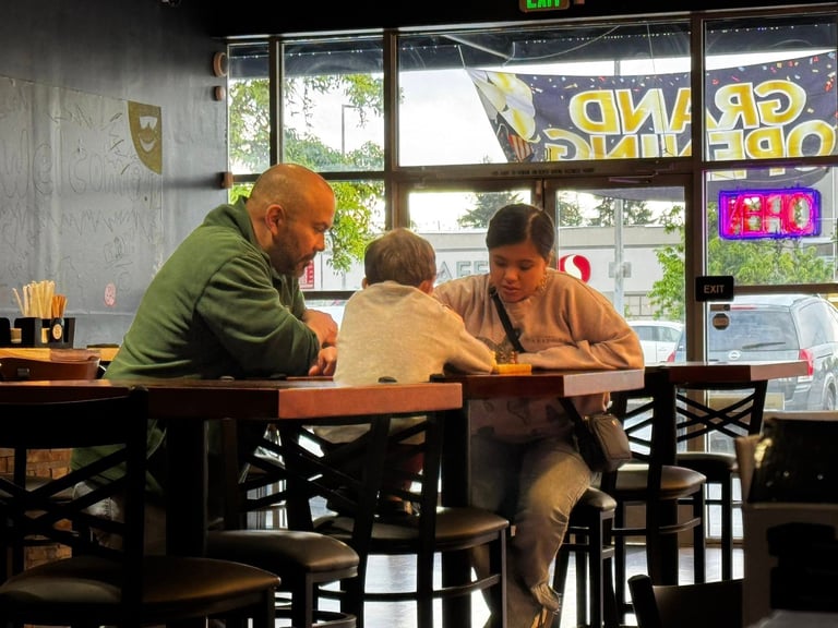 Three people sitting at a wooden table in a cafe near large windows with outdoor views and neon signs