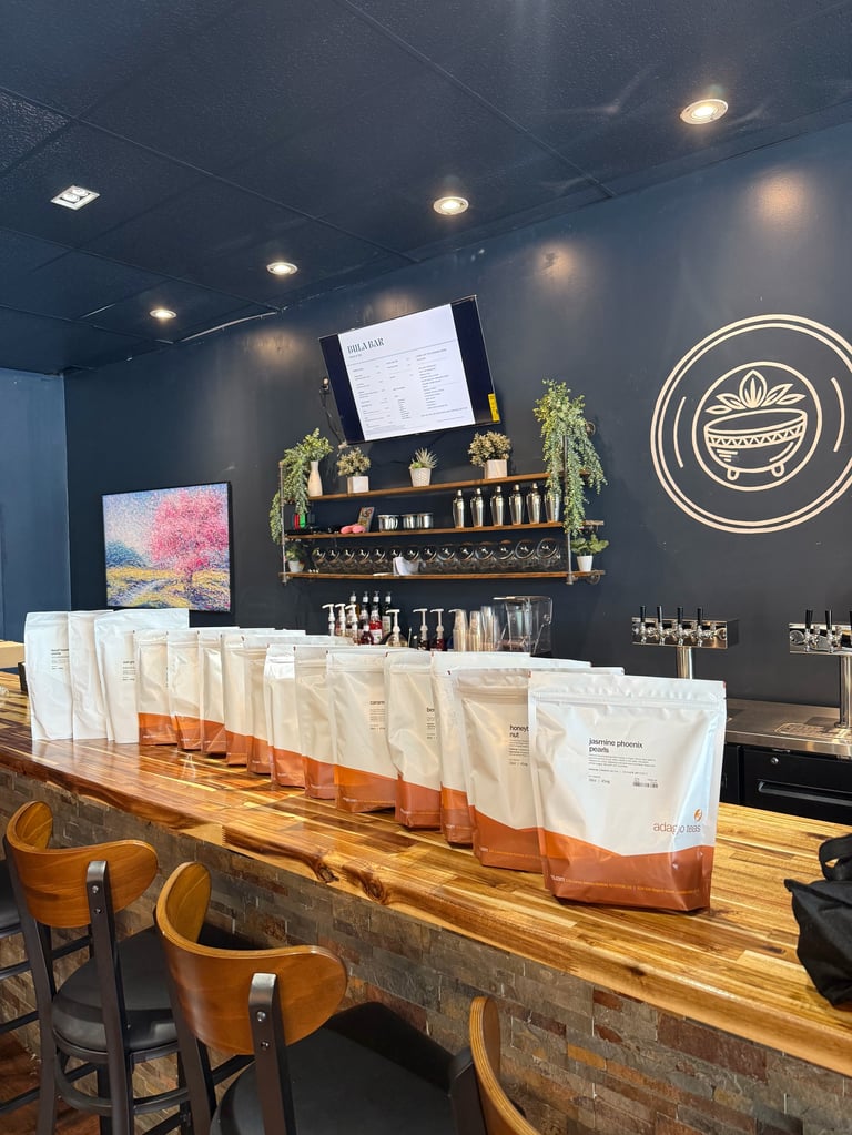 Modern cafe counter with white and orange coffee bags displayed on wood surface, dark blue wall, shelving with plants, and neon logo sign