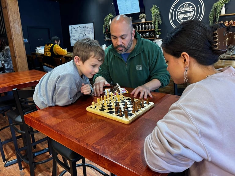 Adult man in green jacket playing chess with young boy and woman at wooden table in indoor setting
