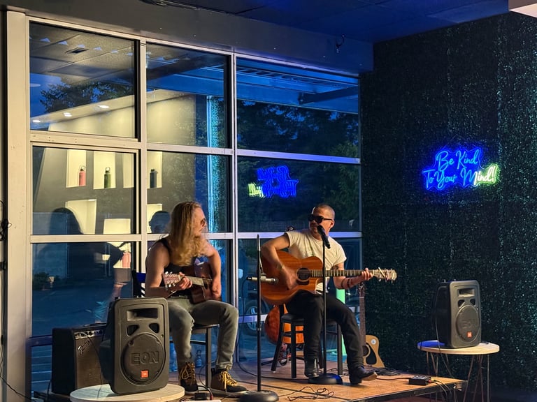 Two musicians perform indoors at night. A woman in the foreground plays a stringed instrument while a man plays acoustic guitar. Blue neon signage glows on the dark wall behind them.
