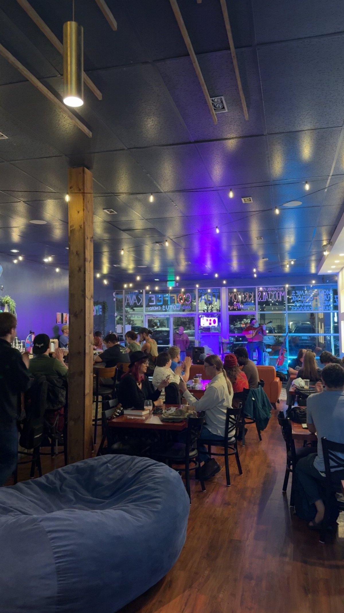 Modern bar or nightclub interior with purple and blue lighting, wooden floor, seated patrons, bar shelving, and industrial ceiling elements.