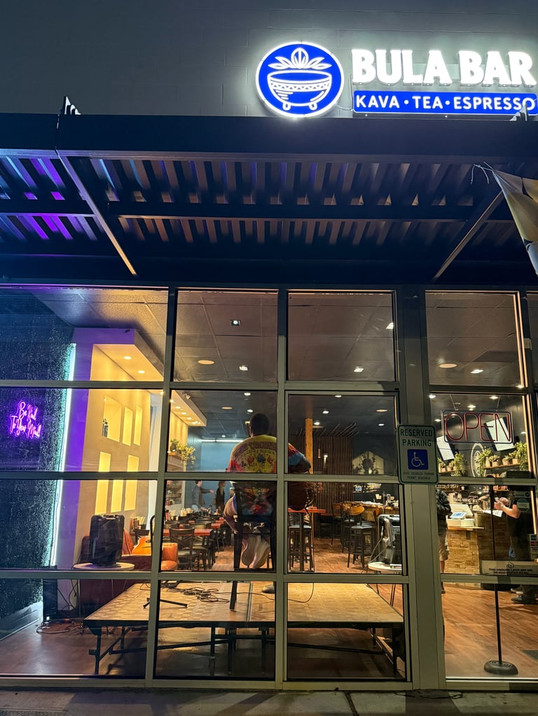 Bula Bar storefront at night with illuminated neon sign advertising kava, tea, and espresso. Glass facade shows interior seating area.