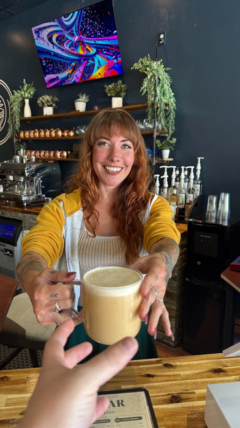Barista with red hair wearing yellow cardigan smiling while handing a cream-colored coffee drink across a wooden counter