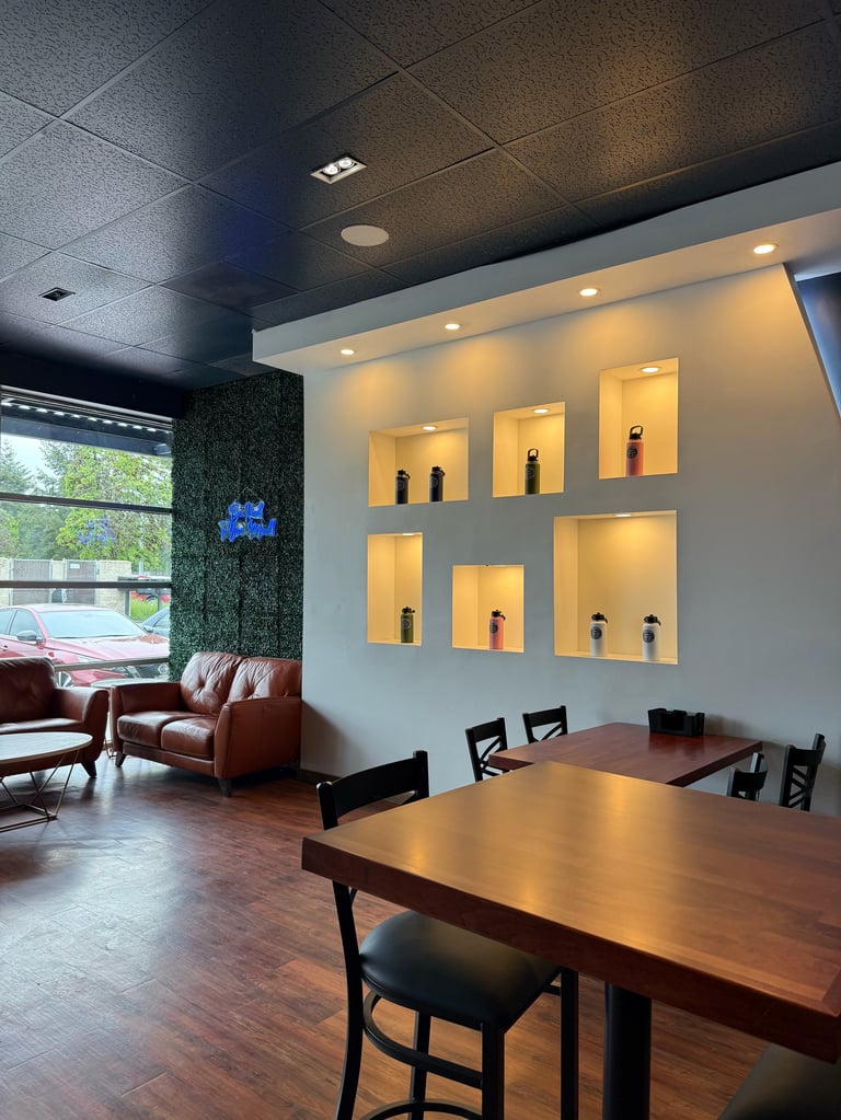 Modern office lounge with dining table, black chairs, brown leather sofa, illuminated wall shelves, and green plant accent wall