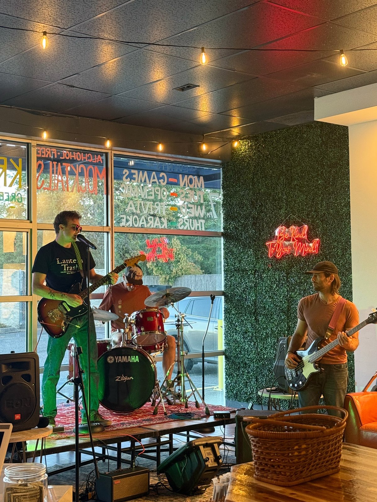 Live band performing indoors with guitarist, drummer, and bass player on stage with neon sign and green moss wall backdrop