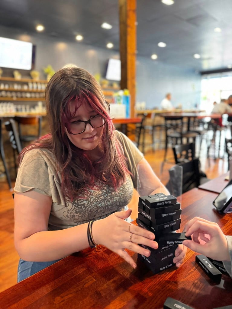 Woman with purple-tinted hair and glasses examines a stack of black cards or devices at a wooden counter in a bright cafe setting.