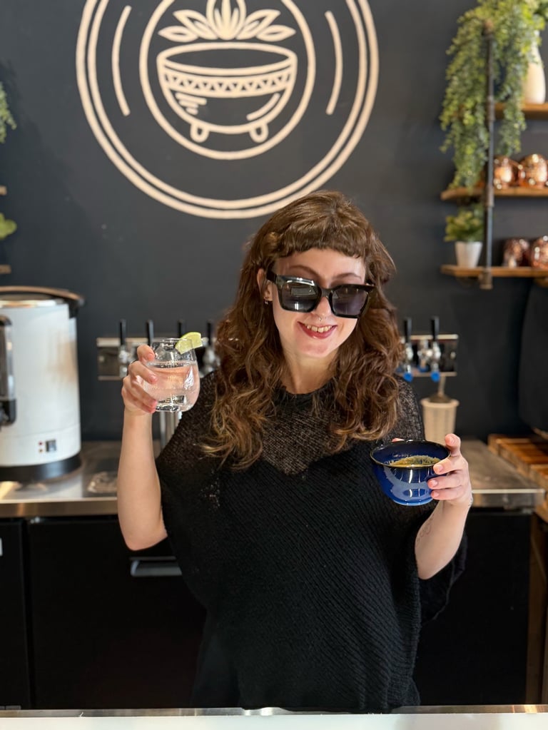 Woman smiling at a bar holding a cocktail and coffee cup, with a cauldron logo on the dark wall behind her