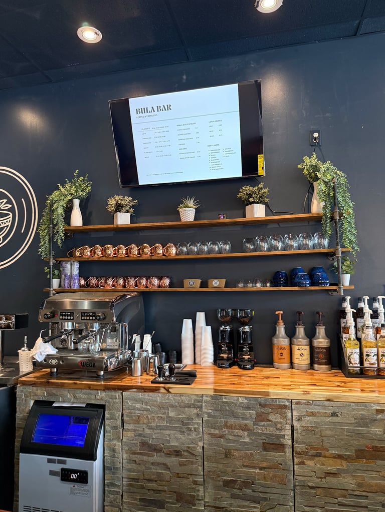 Modern bar counter with wooden shelves displaying glasses and bottles, menu screen above, espresso machine, and hanging greenery on dark wall