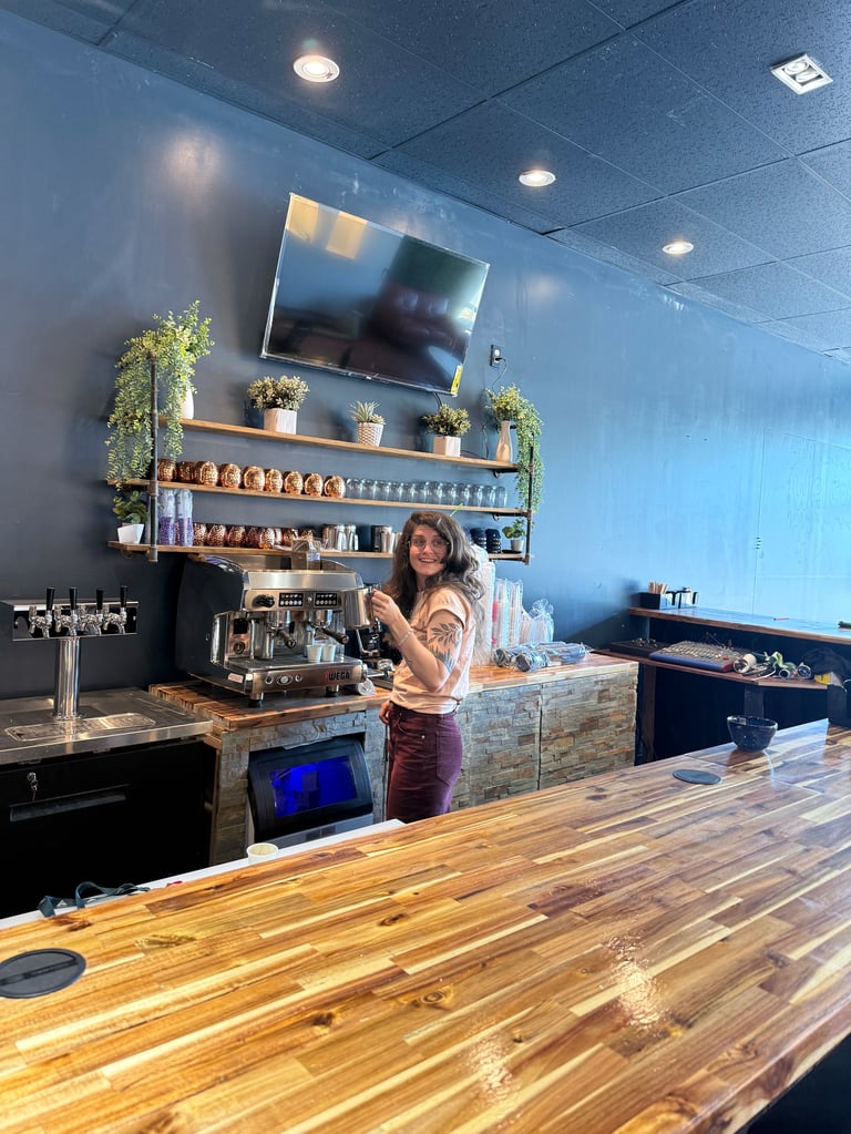 Barista preparing drinks behind wooden counter in modern blue-walled cafe with TV and potted plants on shelving above