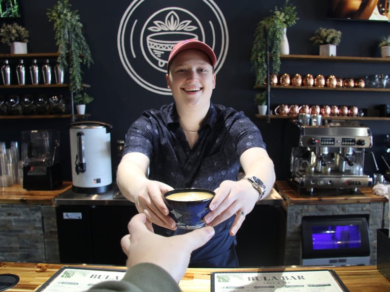 Barista in red cap and blue shirt serving a cup at a coffee shop counter with shelves and neon sign behind