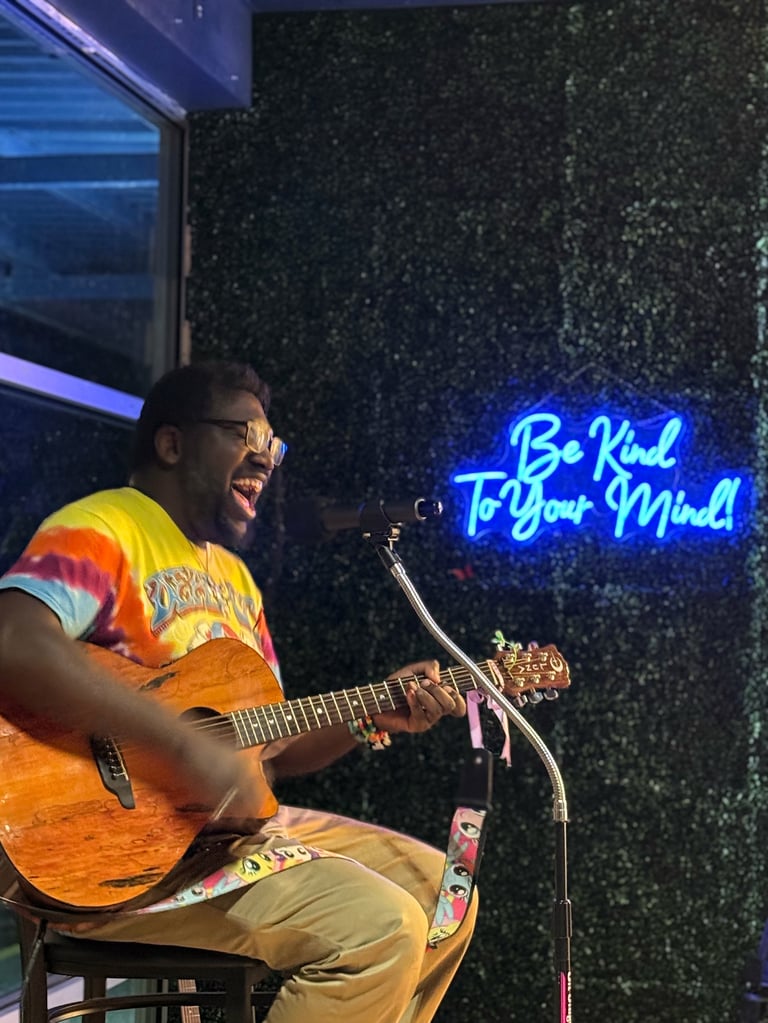 Man playing acoustic guitar on stage wearing yellow shirt, with blue neon sign reading Be Kind To Your Mind in background