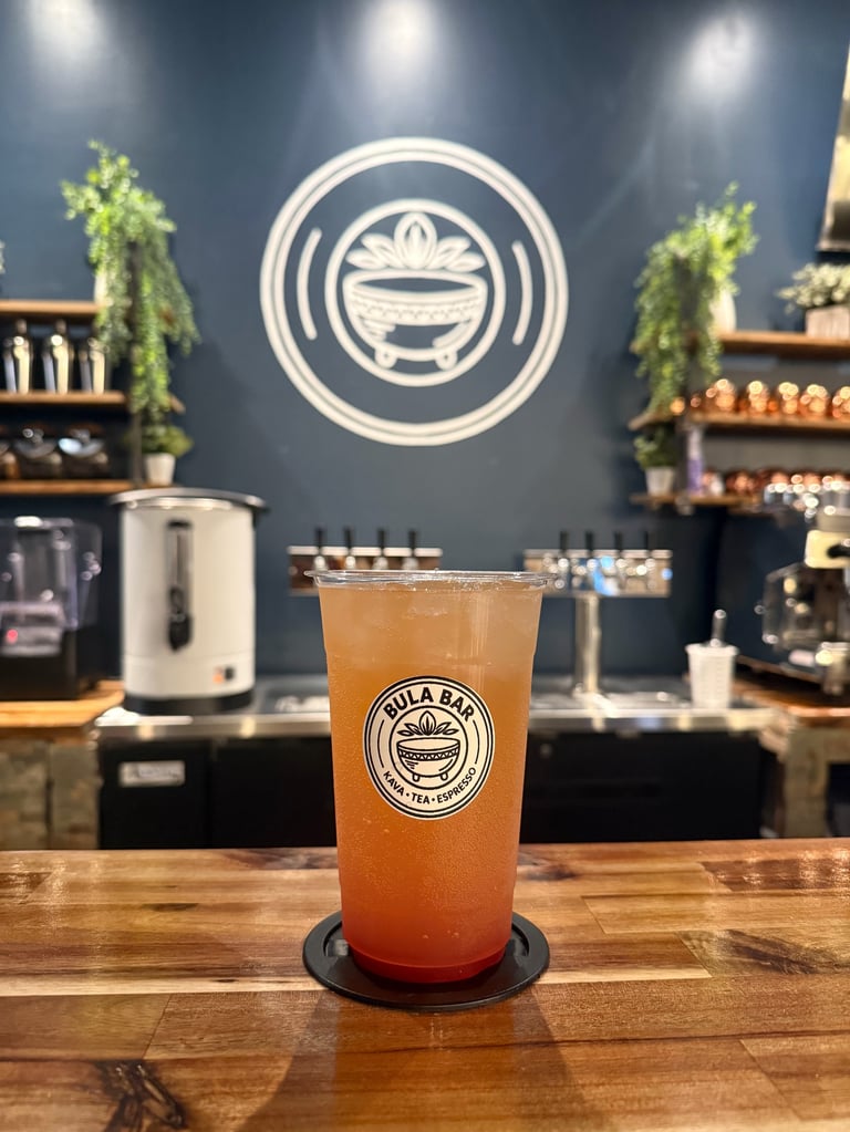 Iced beverage with Bula Bai logo on wooden counter in modern blue cafe with hanging plants and branded circular logo on wall
