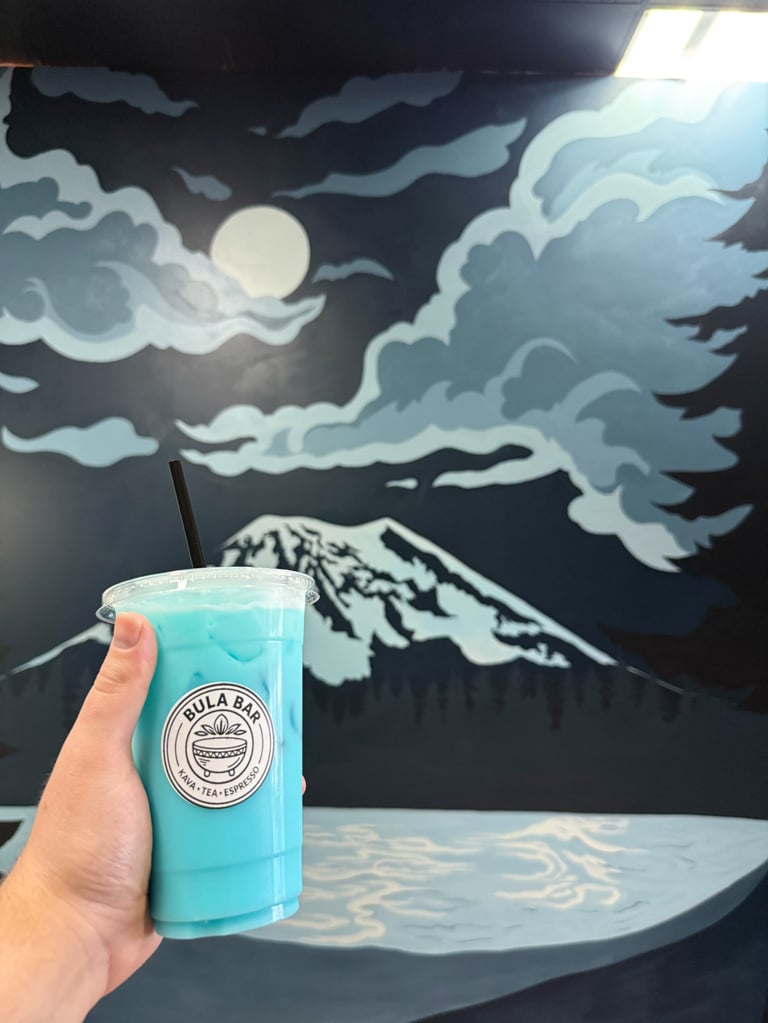 Hand holding a turquoise beverage cup with a circular logo in front of a mural featuring a moon, clouds, and snow-capped mountain