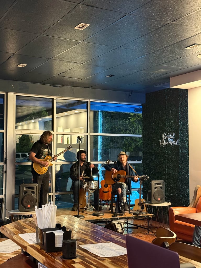 Live music performance with four musicians playing guitar, violin, cello, and drums in a modern indoor venue with large windows.