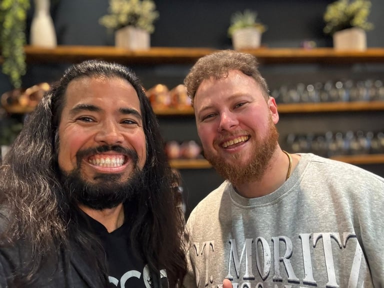Two men smiling at camera indoors with shelves of plants and bottles visible in background