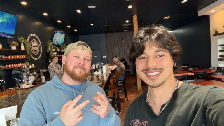 Two men smile at the camera inside a bar or restaurant with dim lighting and beer signage visible in the background