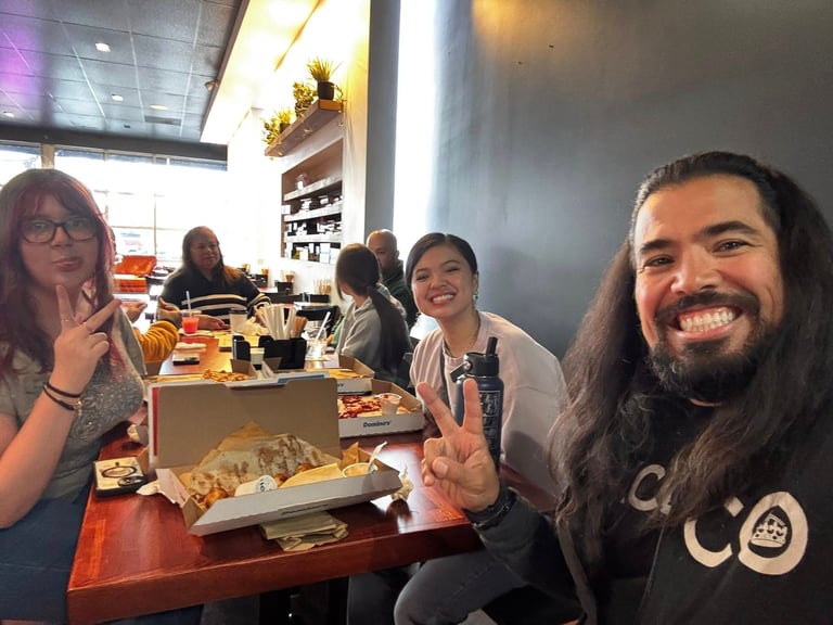 Group of people enjoying food and drinks at a casual restaurant or dining venue, smiling at the camera while seated at a table.